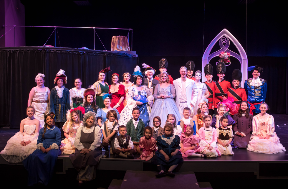 A diverse cast in colorful costumes poses on stage after a theatrical performance.